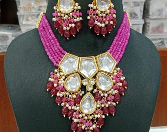 May include: A magenta beaded necklace and matching earrings set. The necklace features large, clear, geometric-shaped stones with gold-coloured metal accents, and dangling red and white beads. The earrings mirror the necklace design, creating a cohesive jewellery set.