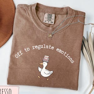 Può includere: T-shirt color espresso con la scritta "Off to regulate emotions" e un'anatra dei cartoni animati con libri in testa. Una collana d'argento con un ciondolo è appoggiata sulla maglietta. La maglietta è piegata su una superficie bianca.
