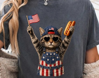 May include: A charcoal gray t-shirt featuring a graphic of a tabby cat wearing heart-shaped sunglasses, a blue baseball cap, and denim overalls with a star and stripe pattern. The cat holds a small American flag and a hot dog in its paws.