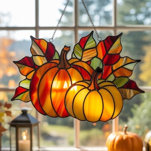 May include: Stained glass pumpkin and leaf decoration. The pumpkins are orange and yellow with black outlines. The leaves are red, orange, and green. The decoration hangs from a chain and is displayed in front of a window.