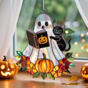 May include: A Halloween-themed stained glass window hanging featuring a ghost reading a book with a jack-o'-lantern on the cover, a black cat, and a pumpkin. The design includes colorful flowers and pumpkins.