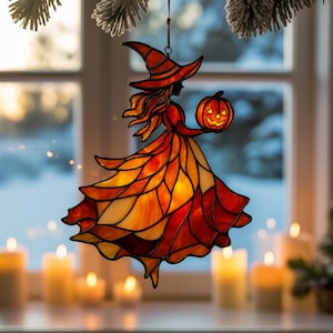 Può includere: Decorazione di Halloween in vetro colorato raffigurante una strega che tiene in mano una zucca intagliata. La strega indossa un cappello a punta e un abito fluente nei toni dell'arancione, del giallo e del rosso. La decorazione è appesa a un filo viola.