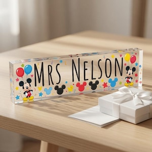 May include: A rectangular clear acrylic desk sign featuring the black text MRS. NELSON, decorated with colourful Mickey Mouse silhouettes, balloons, and star shapes. A small white gift box with a white ribbon sits in front of the sign.