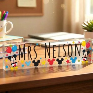 May include: A clear rectangular acrylic desk sign featuring the black text "MRS NELSON" in the centre. The design includes colourful illustrations of Mickey Mouse, red and blue balloons, and various small stars and circles in primary colours.