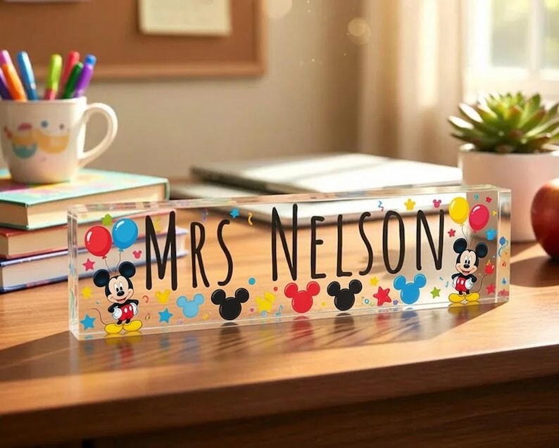May include: A clear rectangular acrylic desk sign featuring the black text "MRS NELSON" in the centre. The design includes colourful illustrations of Mickey Mouse, red and blue balloons, and various small stars and circles in primary colours.