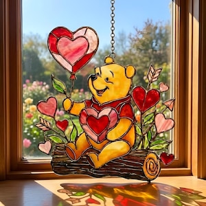 May include: A stained glass artwork featuring Winnie the Pooh holding a heart-shaped balloon and surrounded by heart-shaped accents. The piece shows a yellow bear in a red shirt, sitting on a log. It is suspended by a chain and set against a window.