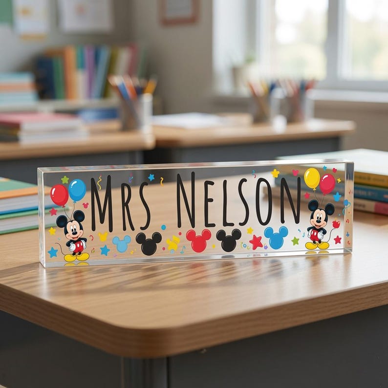 May include: A rectangular clear acrylic desk sign featuring the black text MRS NELSON in the centre. The design includes two illustrations of Mickey Mouse, colourful balloons, and small star and Mickey-shaped icons in red, blue, and yellow.