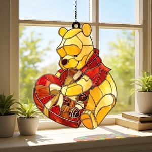 May include: Stained glass window decoration of a yellow bear holding a heart-shaped chocolate box. The bear wears a red scarf and the box has a white bow. The window art hangs from a chain in front of a window with plants and books.