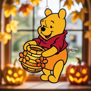 Könnte beinhalten: Gelbe Winnie-the-Pooh-Figur mit rotem Hemd, die einen Honigtopf hält. Das Bild ist eine Fensterdekoration mit dem Text "Winnie the Pooh". Zwei beleuchtete Kürbisse stehen auf der Fensterbank.