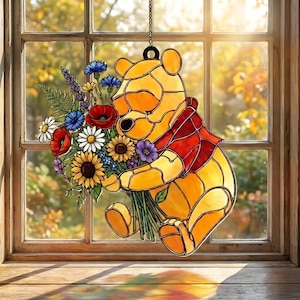 Puede incluir: Decoración de ventana de vidriera con un oso amarillo sosteniendo un ramo de flores coloridas. El oso lleva una bufanda roja y está suspendido por una cadena. El fondo muestra una ventana con vistas a una escena soleada al aire libre.