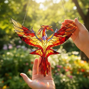 May include: Stained glass phoenix suncatcher with a vibrant color palette of red, orange, and yellow. The bird is suspended by a chain, showcasing its wings spread wide. The suncatcher is a decorative item, perfect for adding a touch of color and artistry to any space.