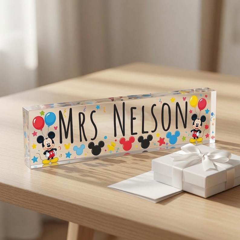 May include: A rectangular clear acrylic desk sign featuring the black text MRS. NELSON, decorated with colourful Mickey Mouse silhouettes, balloons, and star shapes. A small white gift box with a white ribbon sits in front of the sign.