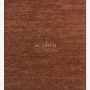 Pode incluir: Um tapete retangular castanho com uma superfície texturizada. O tapete tem uma cor sólida e um design simples. O texto "Indianknots" está no centro do tapete.