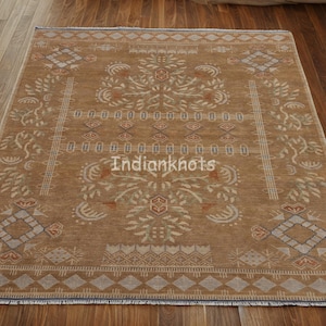 May include: A rectangular, tan-colored rug with an intricate, symmetrical design. The rug features floral and geometric patterns in shades of beige, brown, and rust. The text "Indianknots" is visible on the rug. The rug is placed on a wooden floor.