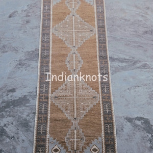 May include: A rectangular woven rug with a central diamond pattern in shades of brown, beige, and grey. The rug features a border with a repeating geometric design and the text "Indianknots". The rug is designed for indoor use.