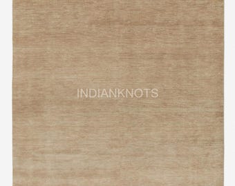 Puede incluir: Una alfombra rectangular de color beige claro sólido con una textura horizontal sutil en toda la superficie. La palabra INDIANKNOTS está impresa en una fuente clara y neutra en el centro de la alfombra, que presenta un borde con flecos cortos arriba.