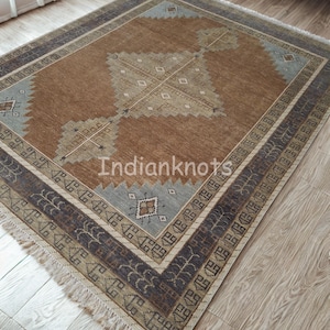 May include: A rectangular brown rug with a central geometric design in beige and light blue. The rug has a patterned border in shades of brown and blue, with a fringe along the edges. The rug is placed on a wooden floor.