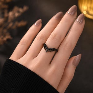 Puede incluir: Un anillo negro en forma de V que presenta un patrón delicado e intrincado similar a una telaraña en la banda. El metal tiene un acabado oscuro y mate, y el diseño forma una punta en chevron que se asienta sobre el dedo, mostrando un trabajo detallado.