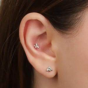 Puede incluir: Primer plano de una oreja con dos pendientes pequeños plateados con forma de abeja. Un pendiente está en el hélix y el otro en el lóbulo. Los pendientes tienen alas y segmentos corporales detallados.