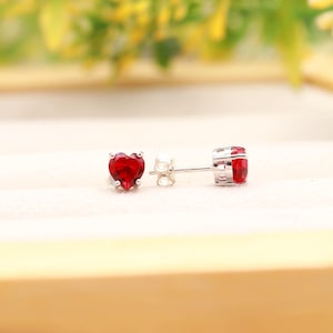Puede incluir: Un par de pendientes de botón con gemas rojas en forma de corazón engastadas en metal plateado. Cada pendiente tiene un engaste de garras que sujeta la piedra roja facetada, y se ve un cierre de presión plateado junto a uno de ellos.