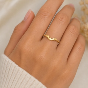 Puede incluir: Un anillo fino de oro con un pequeño adorno central en forma de murciélago. La banda es lisa y pulida, con la silueta del murciélago colocada horizontalmente sobre el dedo. El metal tiene un acabado en oro amarillo brillante y reflectante.