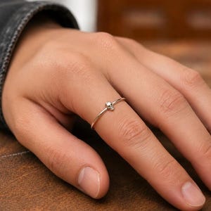 May include: A thin, polished silver-tone ring featuring a small, detailed skull charm centred on the band. The metal has a smooth, reflective finish, and the design consists of a simple, slender circular wire that encircles the finger.