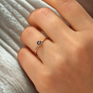 Puede incluir: Un delicado anillo de oro con un pequeño dije redondo en el centro, con un centro azul oscuro y un contorno blanco, que representa un ojo. El anillo se lleva en un dedo, mostrando su diseño minimalista y elegante. La banda del anillo es fina y pulida.