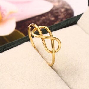 Puede incluir: Un anillo de oro pulido que presenta un diseño de símbolo de infinito continuo. La banda es delgada y lisa, formando un bucle en forma de ocho en la parte superior. El metal tiene un acabado amarillo brillante y reflectante que capta la luz.