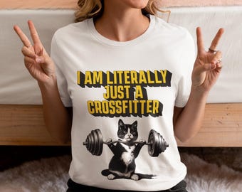 Op de afbeelding: Wit T-shirt met de tekst "I AM LITERALLY JUST A CROSSFITTER" in geel en zwart. Een zwart-witte kattenafbeelding tilt een halter. Het shirt heeft een ronde hals en lijkt van katoen te zijn.