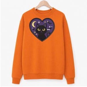 May include: An orange long-sleeve sweatshirt featuring a central heart-shaped graphic. Inside the purple heart is a black cat with yellow eyes, a yellow crescent moon, small white stars, and silhouettes of flying bats against a dark background.
