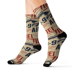 May include: Tan crew socks featuring a repeating graphic pattern of AIM-9X missiles in blue and white, with bold red text reading AIM-9 and the phrase ALL-ASPECT AIR SUPERIORITY MISSILE. The socks have solid black toe and heel sections.