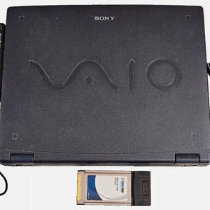 Peut inclure: Un ordinateur portable Sony VAIO noir avec un adaptateur secteur et une carte PC. Le logo VAIO est en relief sur le couvercle. L'adaptateur secteur est noir. La carte PC est argentée et noire.