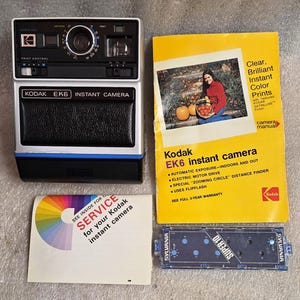 Puede incluir: Una cámara instantánea Kodak EK6 vintage con cuerpo negro y plateado, detalles azules y el texto "KODAK EK6 INSTANT CAMERA". También se incluyen un manual de la cámara, una guía de servicio y un flash Sylvania Super 10.