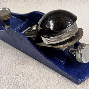 May include: A small, blue hand plane with a black cap and silver adjustment knobs. The plane's body is a deep blue, and the blade is visible within the frame. The knobs and adjustment wheels are silver. This woodworking tool is designed for precision and detail work.