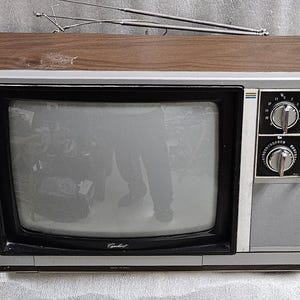 Puede incluir: Un televisor portátil vintage con una parte superior de grano de madera y carcasa plateada. El frente presenta una pantalla negra, una rejilla de altavoz y dos perillas circulares etiquetadas como VHF y UHF. Una antena telescópica está arriba.