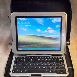 May include: A silver HP tablet computer with a black keyboard base, featuring a screen displaying the classic Windows XP Bliss wallpaper. The device is positioned inside a black zippered carrying case, showing a stylus docked at the top.