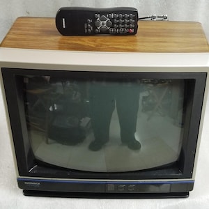 Könnte beinhalten: Ein Vintage-Magnavox-Fernseher mit einer Oberseite in Holzoptik und einem silber-schwarzen Rahmen. Der Fernseher verfügt über einen schwarzen Bildschirm und eine schwarze Fernbedienung, die oben aufliegt. Der Markenname Magnavox steht in Weiß vorne.