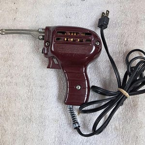 May include: A maroon-colored electric soldering gun with a pistol-grip handle, a metal tip, and a black power cord. The tool features a trigger mechanism, ventilation slots on the side, and a coiled strain relief spring where the cord enters the base.