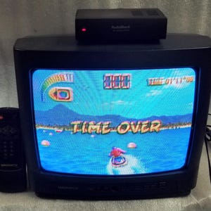 May include: A black Magnavox CRT television displaying a jet ski video game with the text TIME OVER on the screen. A black RadioShack RF modulator with a red light sits on top of the television, and a black Magnavox remote control is positioned to the left.