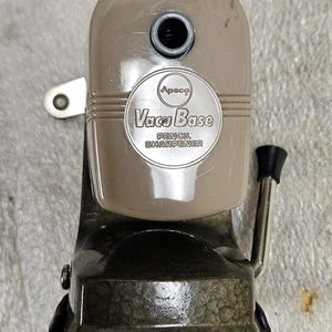 May include: A vintage Apsco Vacu Base pencil sharpener with a beige upper housing and a dark green hammered metal base. The front features a circular silver plate with the text Apsco Vacu Base PENCIL SHARPENER. It includes a side crank handle.