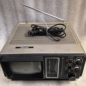Könnte beinhalten: Ein silber-schwarzer tragbarer Bohsei-Fernseher mit eingebauter Antenne, einem oben aufgewickelten schwarzen Netzkabel, einem kleinen Bildschirm, einem vertikalen Kanal-Tuner und runden Drehknöpfen für Strom, Lautstärke und Sendersuche.