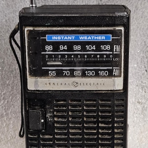 Peut inclure: Radio portable noire General Electric avec une antenne télescopique. La radio a un cadran de réglage avec les fréquences FM et AM, et le texte "INSTANT WEATHER" est affiché au-dessus du cadran. La radio a une sangle noire.