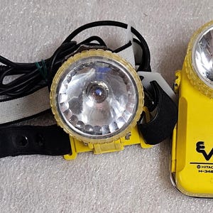 May include: A yellow Hitachi H-3480 headlamp with a large, round light and a black strap. The headlamp has a black cord and a white and black head strap. The headlamp is designed for hands-free illumination.