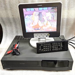 Könnte beinhalten: Ein schwarzer Mitsubishi Videorekorder mit Twin Digital Laufwerk, kombiniert mit einem Casio Fernseher, der einen Zeichentrickfilm zeigt. Das Set enthält eine schwarze Fernbedienung mit vielen Tasten und rot-weiße RCA-Audiokabel.