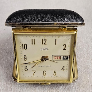 May include: A vintage gold-tone Bradley Day & Date alarm clock with a cream-colored face. The clock displays the time, day, and date, with a black leatherette cover. The clock is marked "Japan".
