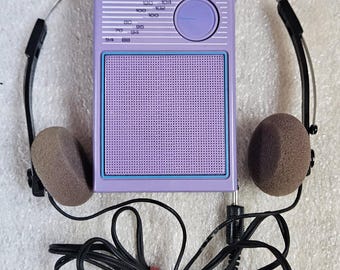 Op de afbeelding: Een lavendelkleurige draagbare AM/FM-radio van Soundesign met een rechthoekig luidsprekerrooster, een ronde afstemknop en een frequentieschaal. De radio is verbonden met een lichtgewicht hoofdtelefoon met bruine schuimrubberen oorkussens.