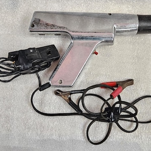 Puede incluir: Una lámpara estroboscópica automotriz plateada con un mango negro y un botón rojo. Está conectada a una caja negra y cables negros con pinzas rojas y negras. La herramienta está diseñada para uso automotriz.