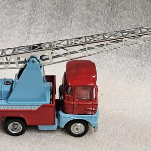 May include: A toy crane truck in red, light blue, and silver. The truck has a red cab, a light blue body, and a silver crane arm with a hook. The crane arm is extended upwards. The truck has black wheels.