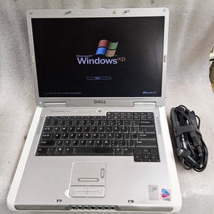 Könnte beinhalten: Ein silber-weißes Dell Inspiron Notebook mit schwarzer Tastatur und Touchpad. Der Bildschirm zeigt das Microsoft Windows XP Logo und den Starttext. Ein schwarzes Netzkabel ist aufgerollt und rechts neben dem Laptop platziert.
