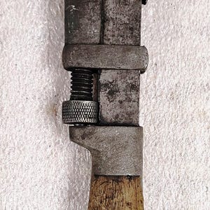 May include: A vintage adjustable wrench featuring a dark, weathered metal head and a sliding jaw mechanism with a knurled adjustment nut. The tool has a worn wooden handle secured with a single metal rivet, showing signs of age and heavy use.